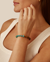 Close-up of a woman's wrist wearing a Chan Luu Unity Bracelet in Turquoise Mix with turquoise and brown beads, her hand touching her neck, against a beige background.