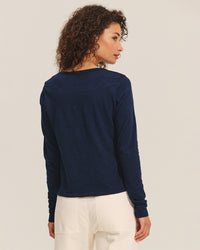 A woman with curly brown hair faces away, looking over her left shoulder, wearing the Becky Long Sleeve Scoop Neck Top in Midnight by Velvet by Graham & Spencer and high-waisted off-white pants against a light background.