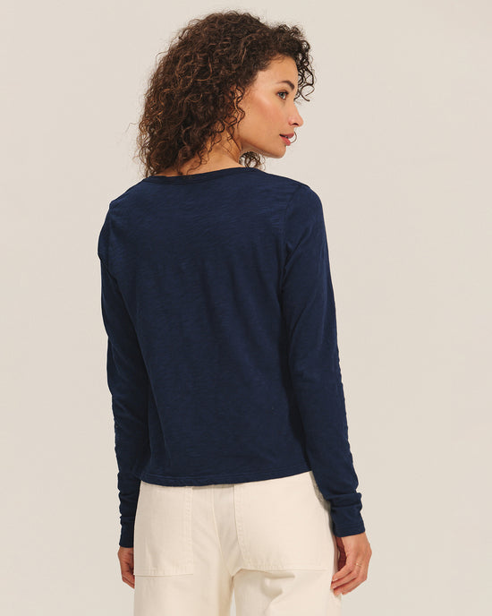 A woman with curly brown hair faces away, looking over her left shoulder, wearing the Becky Long Sleeve Scoop Neck Top in Midnight by Velvet by Graham & Spencer and high-waisted off-white pants against a light background.