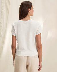 A woman with straight brown hair stands facing away, wearing a Velvet by Graham & Spencer Marika Short Sleeve Crew Neck Crop Top in white and light beige pants against a soft, cream-colored fabric backdrop.