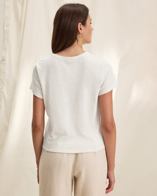 A woman with straight brown hair stands facing away, wearing a Velvet by Graham & Spencer Marika Short Sleeve Crew Neck Crop Top in white and light beige pants against a soft, cream-colored fabric backdrop.