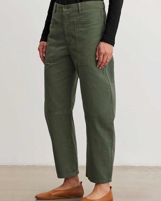 A person stands against a plain white wall wearing Velvet by Graham & Spencer's Brylie Curved Knee Pant in Caper, styled with a black long-sleeve top and brown flats—an effortlessly chic look perfect for casual occasions.