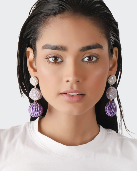 A woman with dark hair wearing white and purple Olivia Dar Venus Earrings in Purple beaded earrings and a white top.
