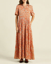A woman wears the Trovata Birds of Paradis Martine Dress in Vermillion Vine, featuring a long, loose fit with an orange floral print on beige, maxi tiered skirt, one hand in a pocket and dark shoes; her head is slightly cropped out.