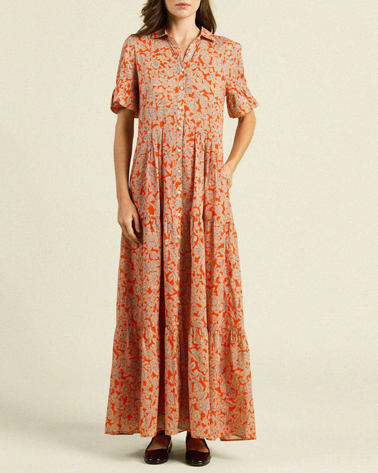 A woman wears the Trovata Birds of Paradis Martine Dress in Vermillion Vine, featuring a long, loose fit with an orange floral print on beige, maxi tiered skirt, one hand in a pocket and dark shoes; her head is slightly cropped out.