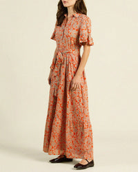 A woman stands wearing the Trovata Birds of Paradis Martine Dress in Vermillion Vine, handmade in California with a red floral pattern on beige, a matching fabric belt, and a flowing maxi-length tiered skirt.