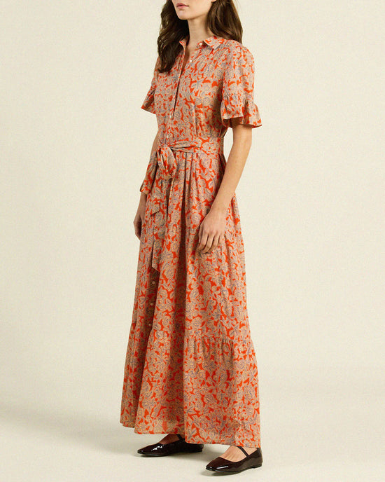 A woman stands wearing the Trovata Birds of Paradis Martine Dress in Vermillion Vine, handmade in California with a red floral pattern on beige, a matching fabric belt, and a flowing maxi-length tiered skirt.