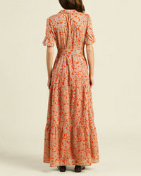 A woman stands facing away, wearing the Martine Dress in Vermillion Vine by Trovata Birds of Paradis—a handmade California maxi with a red and beige floral pattern, short sleeves, fitted waist, and tiered skirt—against a plain light background.
