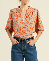 A woman stands against a neutral background with hands in pockets, wearing Trovata Birds of Paradis’s Loren Blouse in Vermillion Vine—a loose-fitting, orange and beige floral puff sleeve blouse—paired with blue high-waisted jeans.