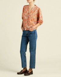 A person stands with hands in pockets, wearing the Loren Blouse in Vermillion Vine by Trovata Birds of Paradis (100% cotton, loose orange floral), blue straight-leg jeans, and brown loafers against a plain light background.