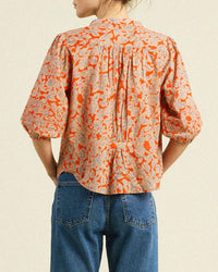 A person is seen from behind wearing the Trovata Birds of Paradis Loren Blouse in Vermillion Vine, a loose-fitting, 100% cotton top with half sleeves, paired with blue jeans against a light background.