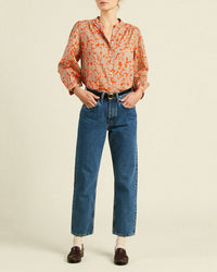 A person stands against a plain background wearing the loose, long-sleeve Clara Blouse in Vermillion Vine by Trovata Birds of Paradis, tucked into high-waisted blue jeans with a black belt, brown loafers, and beige socks.