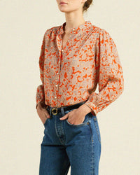 A woman wears the Clara Blouse in Vermillion Vine by Trovata Birds of Paradis—a long-sleeved, floral button-down in orange and beige—with blue jeans and a black belt, standing with hands in her pockets, shown mid-torso up.