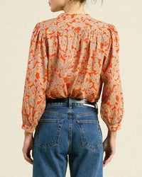 A person wearing the Trovata Birds of Paradis Clara Blouse in Vermillion Vine, a long-sleeved orange floral button-down, tucked into high-waisted blue jeans, shown from the back against a plain light background.