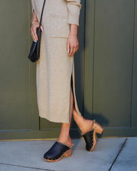 Woman in a beige skirt and sweater set with black clogs featuring the Woodpecker Mas Shearling in Black/Bronze by beek. and carrying a black purse standing against a green wall.