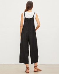 Woman standing with her back to the camera, wearing the Isabel Jumpsuit in Black by Velvet by Graham & Spencer and a white t-shirt, paired with brown sandals.