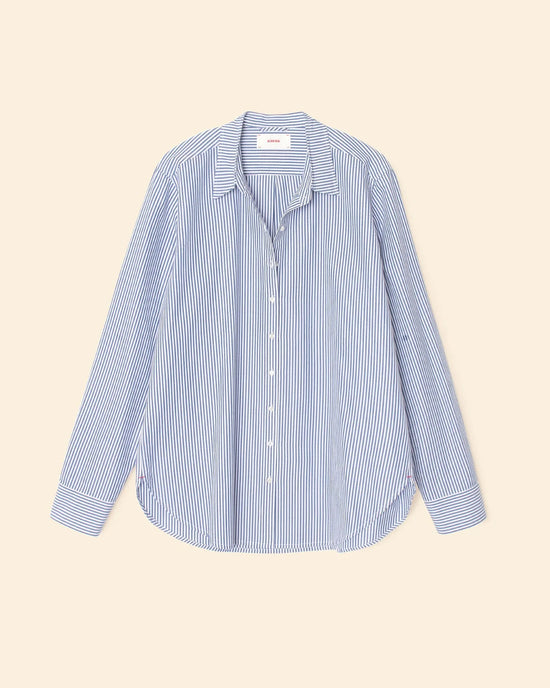 The XiRENA Beau Shirt in True Blue is a 100% cotton, oversized button-down with blue and white vertical stripes, a collar, and relaxed fit, shown laid flat on a beige background.