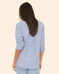 A person with straight, long brown hair is shown from the back wearing the XiRENA Beau Shirt in True Blue and light blue jeans against a plain beige background.