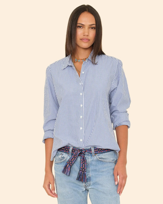 A woman with straight dark hair wears the XiRENA Beau Shirt in True Blue—an oversized blue and white striped button down with rolled-up sleeves—paired with light jeans and a patterned waist scarf, standing against a beige background.