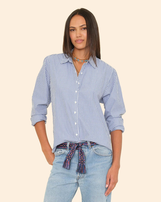 A woman with straight, dark hair wears an oversized XiRENA Beau Shirt in True Blue, featuring blue and white stripes, styled with light jeans, a colorful belt, and layered necklaces against a neutral background.