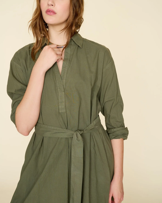 A person wearing a green XiRENA Hope Dress in Ash Green with a tied waistband, posing with one hand near the collar.