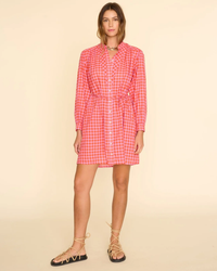 Woman in a red and white checkered XiRENA Winnie Dress in Orange Pink with long sleeves, standing against a beige background.