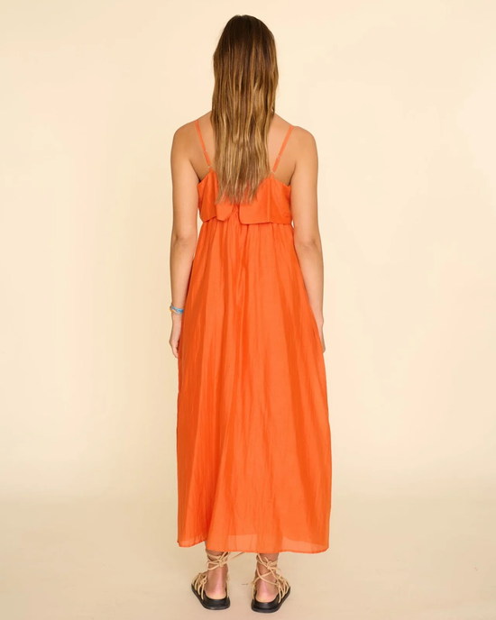 A person with long hair wearing an orange cotton silk voile XiRENA Skyla Dress in Papaya and strappy sandals stands with their back to the camera.