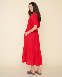 A woman in a red cotton gauze XiRENA Lennox Dress in Real Red with short sleeves, standing sideways to the camera.
