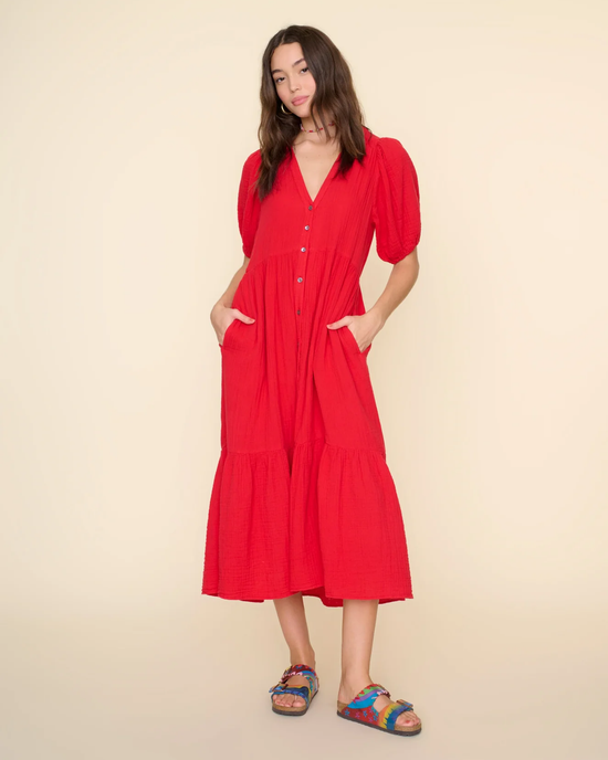Woman posing in a red Cotton Gauze XiRENA Lennox Dress in Real Red with buttons and sandals against a beige background.