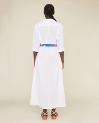 A woman stands with her back to the camera, wearing a white XiRENA Boden dress in maxi length with a colorful belted waist and brown strappy sandals.