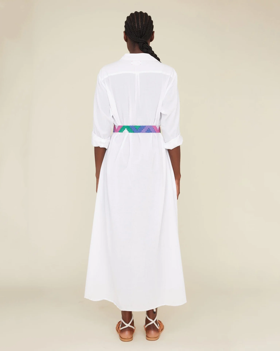 A woman stands with her back to the camera, wearing a white XiRENA Boden dress in maxi length with a colorful belted waist and brown strappy sandals.