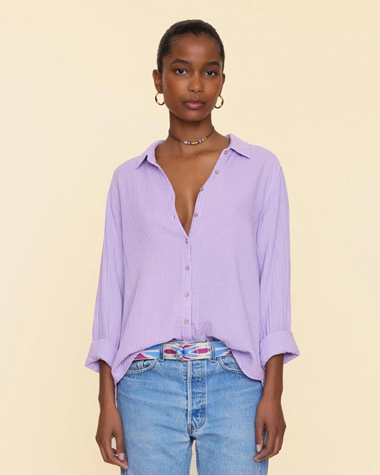 A woman wearing an unbuttoned light purple XiRENA Scout Shirt in Viola, jeans, and simple jewelry stands against a beige background.