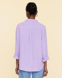 Woman standing with her back to the camera, wearing a light purple XiRENA Scout Shirt in Viola and blue jeans against a beige background.