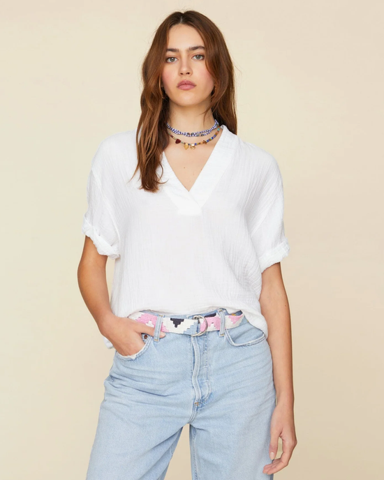 A woman wearing a white v-neck XiRENA Avery Top in cotton gauze, light blue denim jeans, and accessorizing with a beaded choker and a patterned belt.