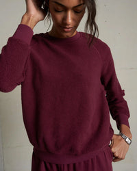 A person wearing the perfectwhitetee Ziggy Inside Out Fleece L/S Shrunken Crew SS in Dark Burgundy stands by a light-colored wall, looking down with one hand on their head and the other on their hip.