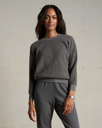 A woman with long dark hair wears the perfectwhitetee Ziggy Inside Out Fleece L/S Shrunken Crew SS in Charcoal and matching gray pants, standing against a plain light-colored wall and smiling softly.