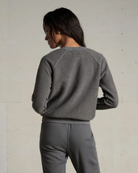 A woman with long dark hair stands facing away, wearing the perfectwhitetee Ziggy Inside Out Fleece L/S Shrunken Crew SS in Charcoal, paired with matching pants against a plain, light-colored wall.