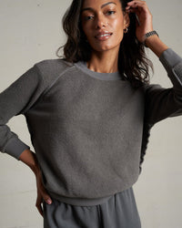A woman with wavy dark hair smiles softly and touches her hair while wearing the perfectwhitetee Ziggy Inside Out Fleece L/S Shrunken Crew SS in Charcoal, posing against a plain light wall.