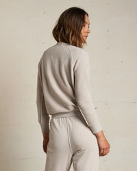 A woman with shoulder-length brown hair stands facing away, wearing the perfectwhitetee Ziggy Inside Out Fleece L/S Shrunken Crew SS in Silver with matching lounge pants, set against a plain off-white wall.