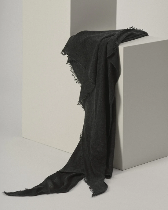 A black, fringed, Grisal Love Cashmere Scarf in Charcoal draped over a gray cube in a minimalist setting.