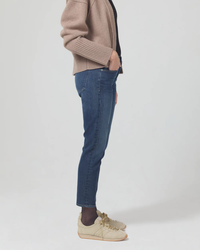 Person standing sideways wearing Citizens Of Humanity Emerson Slim Boyfriend 27" in Trinket jeans and a brown sweater made of organic cotton with one hand in pocket.