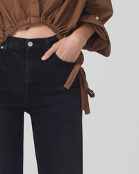 A person wearing a brown jacket and dark jeans with a hand resting on the hip is now in Citizens of Humanity Daphne Crop 26.5" Inseam in Peppercorn Jeans.
