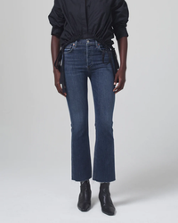 A person stands wearing a black shirt and medium indigo jeans with frayed hems, paired with Citizens of Humanity Isola Cropped Boot in Zip Fly Undercurrent ankle boots.