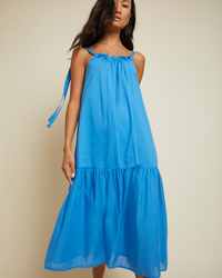 Woman modeling a Nation LTD Sequoia Voluminous Sundress in Lapis, featuring a blue sleeveless summer design with tie straps and a ruffled hem.