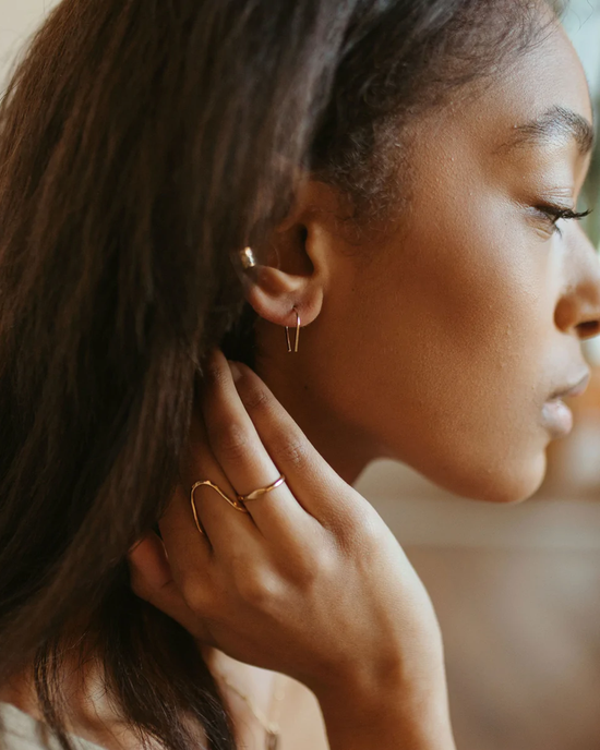 A woman is showcased with a close-up on her profile, subtly revealing elegant Small Horseshoe Earrings in 14K Gold Fill from Token Jewelry and a ring on her finger as she gently touches her neck.