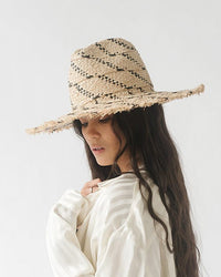 A woman in a white blouse wearing a Gigi Pip Gemma Wide Brim Straw hat in Natural/Black with a fringed edge, looking away from the camera.