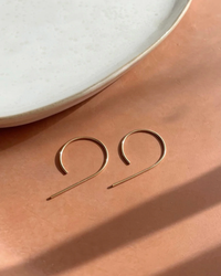 A pair of The Nines Earrings in 14K Gold Fill, handmade in USA, resting on a peach-colored surface next to a white plate from Token Jewelry.