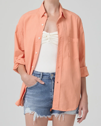 A person modeling a casual outfit featuring an oversized orange button-up Kayla Shirt in Papaya, a white top, and Citizens of Humanity denim shorts.
