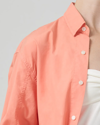 A close-up of a person wearing the Citizens of Humanity Kayla Shirt in Papaya, an oversized coral pink collared shirt with white buttons and a high low hem.