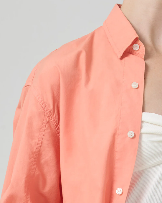A close-up of a person wearing the Citizens of Humanity Kayla Shirt in Papaya, an oversized coral pink collared shirt with white buttons and a high low hem.